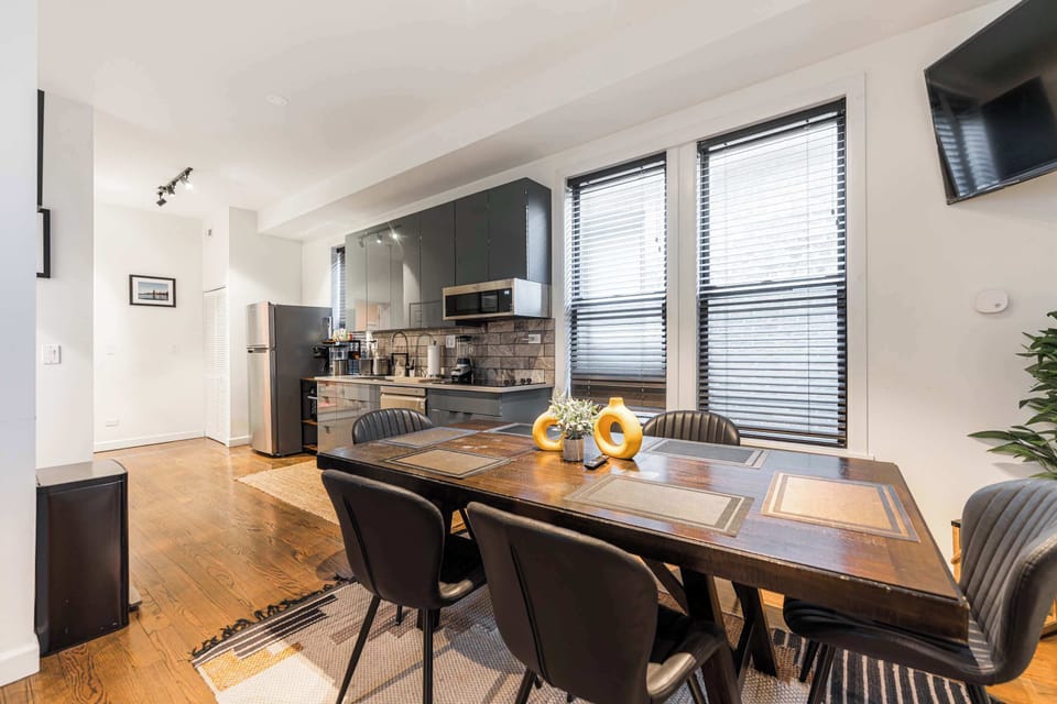 Enjoy meals in this stylish open-concept dining area featuring a rustic wood table and modern seating. Bathed in natural light from large windows, it flows seamlessly into a sleek kitchen with stainless steel appliances and dark cabinetry.