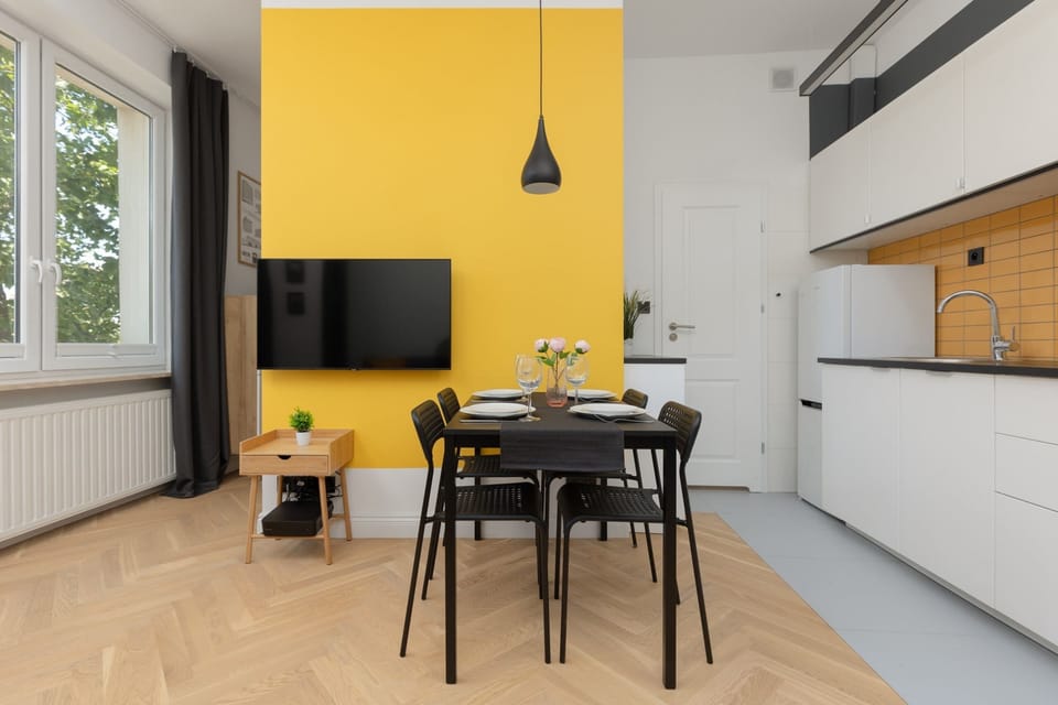 A view of the living room with a dining area. A yellow wall features a mounted TV. The dining table is set with black chairs.
