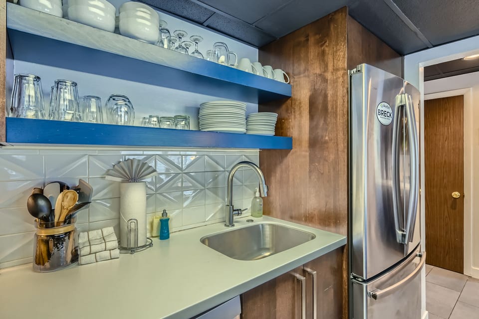 A kitchen area with open shelving holding dishes and glasses, a stainless steel refrigerator, a sink, utensils in a holder, a soap dispenser, and a tiled backsplash.