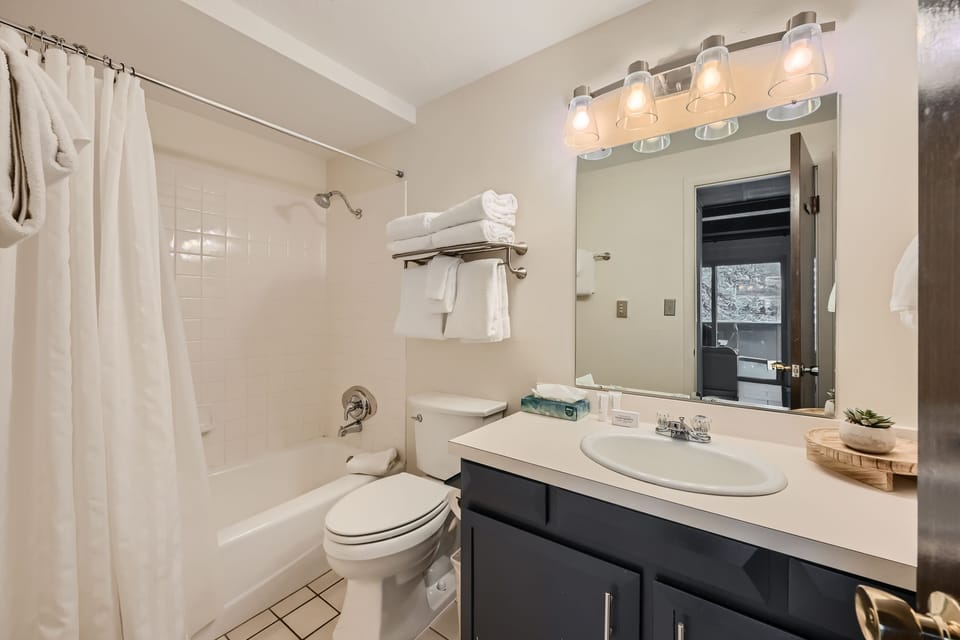 A clean, modern bathroom featuring a bathtub with a shower curtain, a toilet, a sink with a dark vanity, a mirror with light fixtures, and shelves holding neatly folded towels.