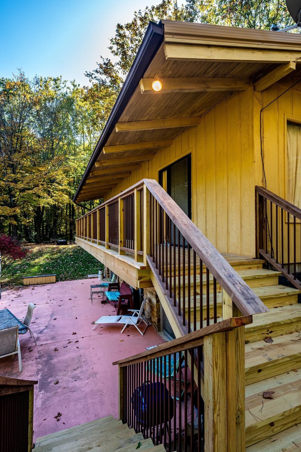 Exterior view of yellow cabin siding with wooden staircase and railing leading to a pink concrete patio with outdoor seating.