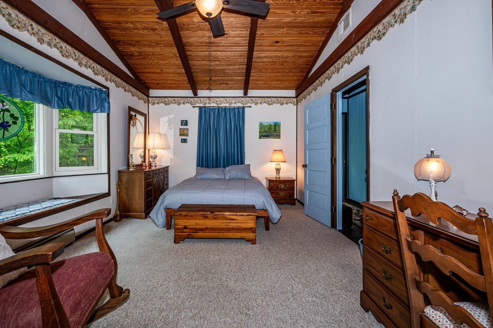 Bedroom with vaulted wood ceiling, floral wallpaper border, blue curtains, wooden furniture, and window seat.