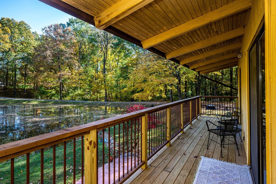 Covered wooden deck with black metal chairs overlooks pond and autumn trees in vibrant fall colors.
