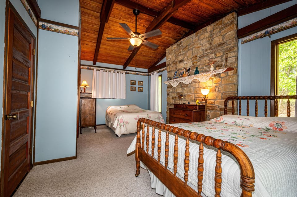 Two wooden spindle beds with floral quilts face a stone accent wall beneath a vaulted wood ceiling with exposed beams.