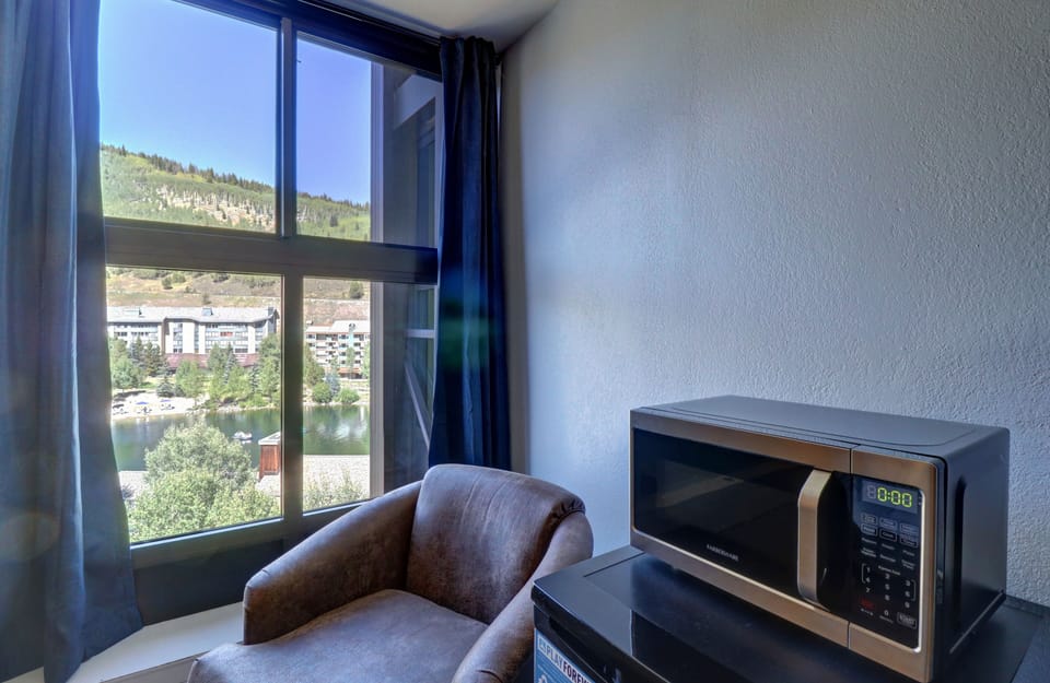 A room with a large window showcasing a hillside view, a brown armchair, and a microwave oven positioned on top of a mini-fridge.