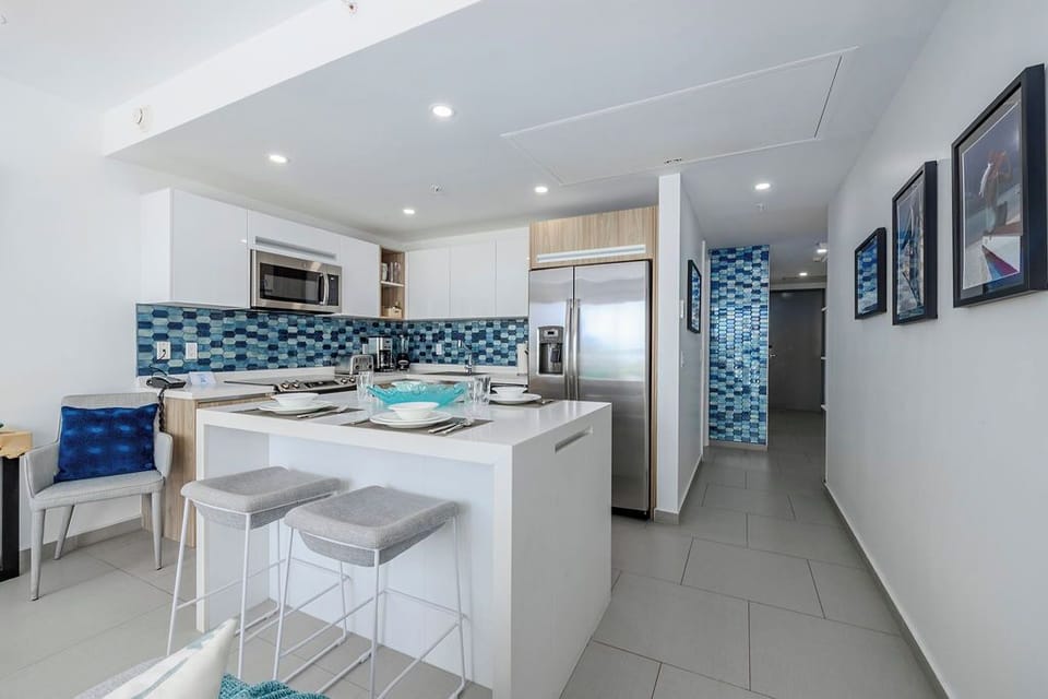 Gather around this sleek modern kitchen where bright white cabinets meet vibrant ocean-blue tile. Your morning coffee becomes a ritual at the island bar, while stainless appliances stand ready for your culinary adventures.