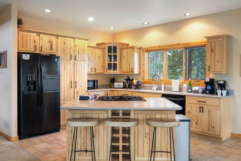 Your fully equipped kitchen features rustic wood cabinetry, modern appliances, and a spacious island with seating—perfect for preparing meals and gathering with loved ones as natural light streams through forest-facing windows.