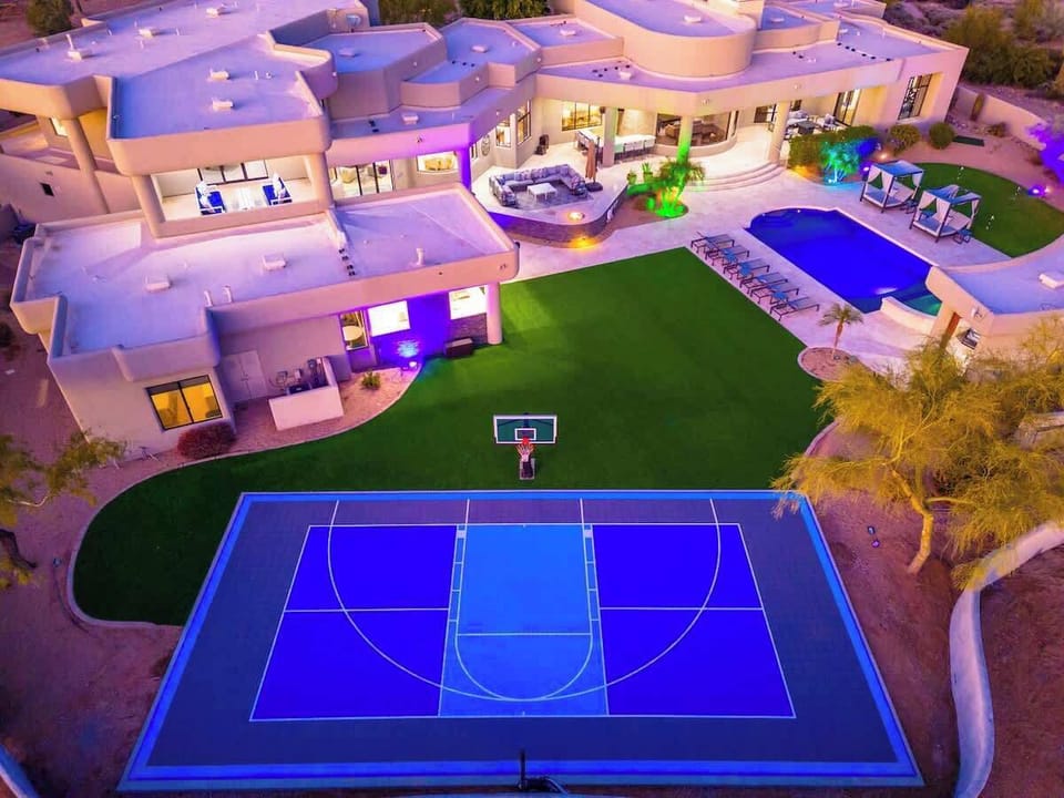 Aerial view of a luxury desert estate featuring a private basketball court, swimming pool, expansive lawns, and modern architecture with dramatic evening lighting—perfect for an unforgettable retreat in the surrounding desert landscape.