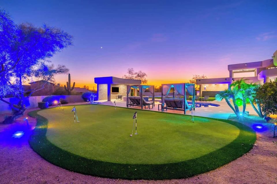 Modern desert retreat featuring a private putting green, illuminated outdoor lounge areas, and stunning evening views of surrounding natural landscape beneath a glowing sunset sky.