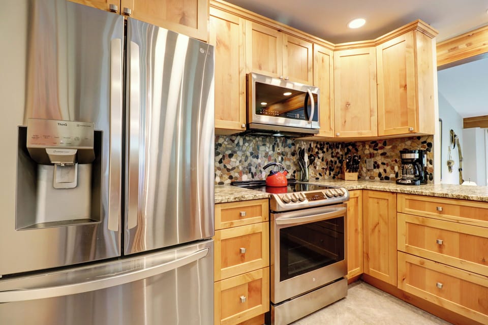 A modern kitchen with stainless steel appliances, wooden cabinets, a mosaic tile backsplash, and a countertop with a coffee maker and a stovetop with a red kettle.