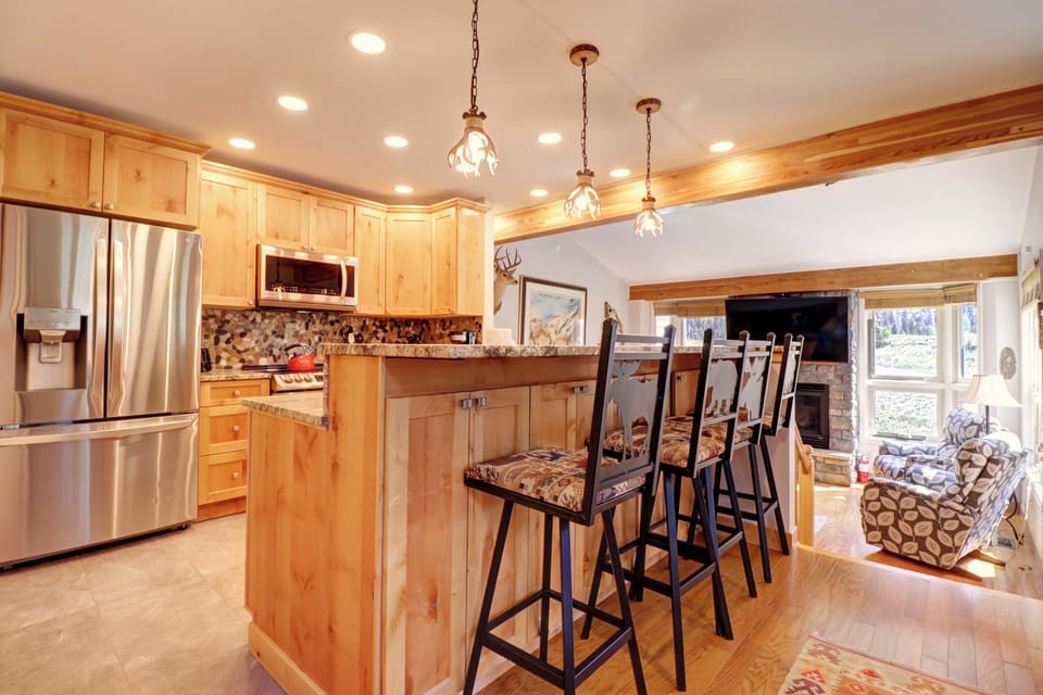 A modern kitchen with wooden cabinetry, stainless steel appliances, a central island with bar stools, hanging pendant lights, and an adjacent open living area with a fireplace and mounted TV.