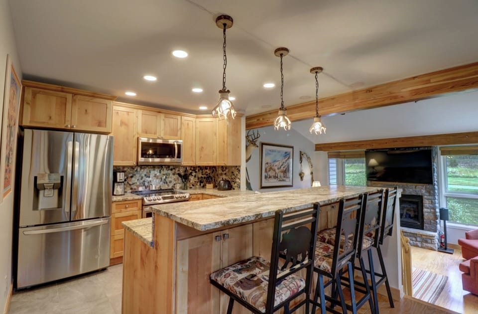 A kitchen with wooden cabinets, a stainless steel refrigerator, and a microwave. A stone backsplash is present above the counter. A long island with bar stools divides the kitchen from the living area.
