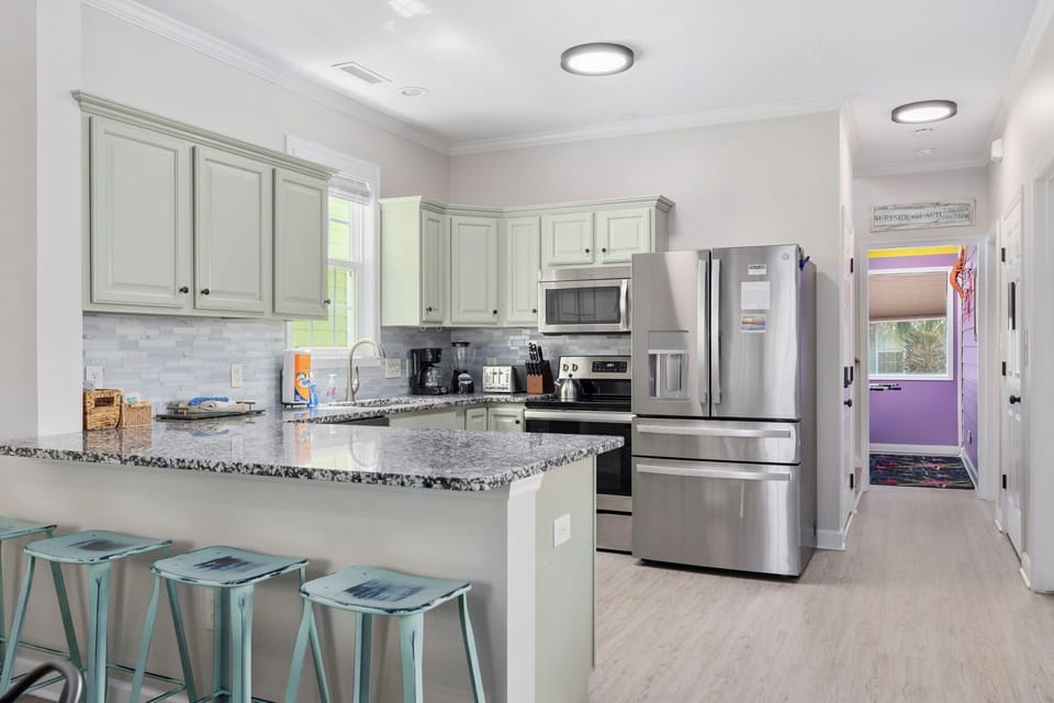 Cook at the granite peninsula with teal bar stools for the sous chefs — sage green cabinets, stainless appliances, and the arcade room visible down the hall.