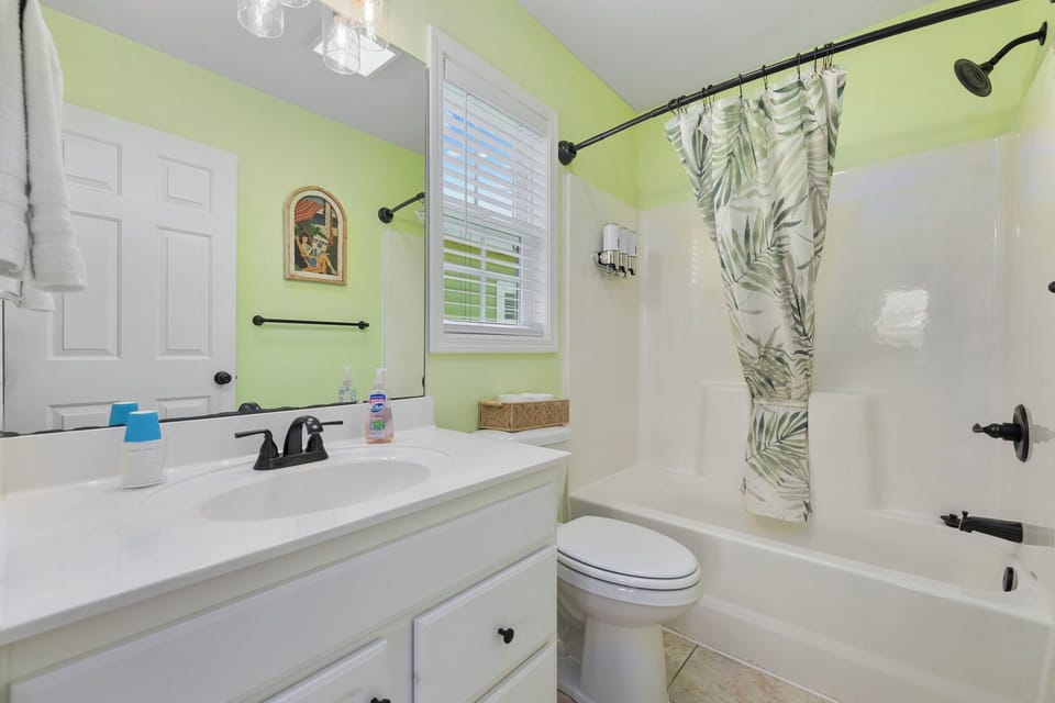 Rinse the salt off in the flamingo room's en-suite — tub and shower combo with a tropical leaf curtain, white vanity, small colorful artwork above.