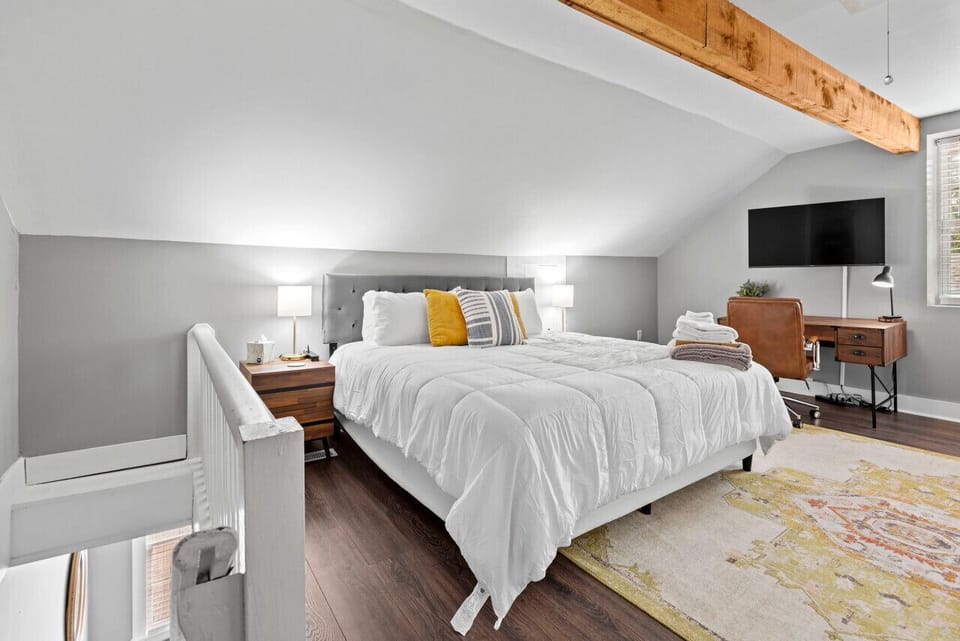 The loft bedroom pairs a bed with exposed wood beams, a wall-mounted TV, a writing desk, and bedside lighting.