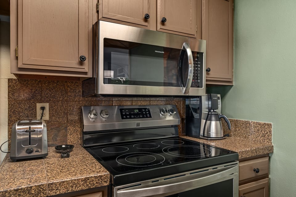 A kitchen with granite countertops features a stainless steel microwave, an electric stove, a coffee maker, and a toaster. The cabinets above and below the counter are light brown.