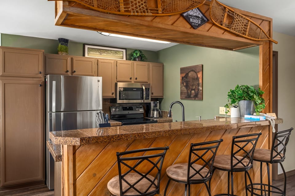 A modern kitchen with light wood cabinets, stainless steel appliances, and a granite countertop bar with four metal chairs. Decor includes a plant, wall art, and a vintage snowshoe hanging above the bar.