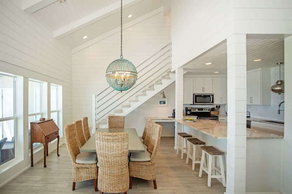 - Gather for meals at a spacious dining table with woven chairs and a beach vibe - Beautiful open kitchen with barstool seating makes cooking and entertaining easy - Stairs lead to even more space, all under bright white paneling and vaulted ceiling