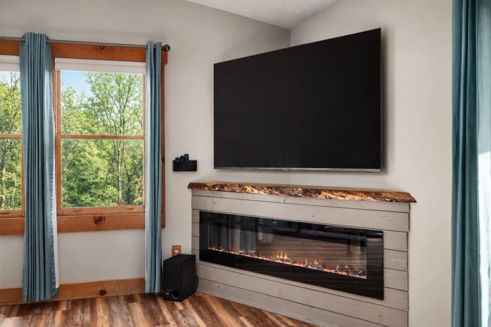 Wall-mounted TV above an electric fireplace with a wood mantel, set in the corner between two windows.