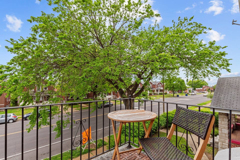 The upper balcony includes a round wood table and woven chair behind a black metal railing, with a leafy daytime view along the residential street.