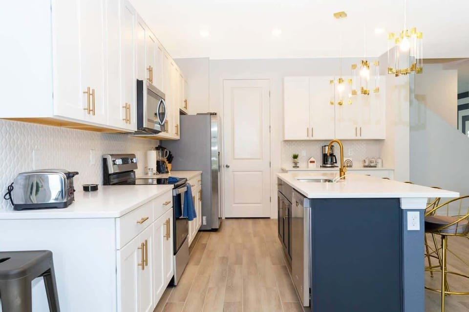 Bright gourmet kitchen-white shaker cabinets, quartz counters, navy island with gold faucet, and stainless appliances under chic pendants. Plenty of space for group cooking!