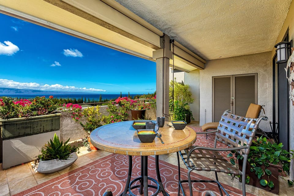 A covered lanai overlooking the Pacific Ocean features vibrant tropical gardens, comfortable seating, and panoramic views of Maui's pristine coastline and neighboring islands under Hawaii's endless blue sky.