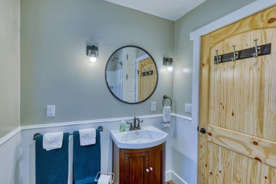 Capture another angle of our bathroom, highlighting the wooden door with hooks, round mirror, sink, and towel bar adorned with towels and face cloths. Every detail adds to the comfort and convenience of this inviting space.