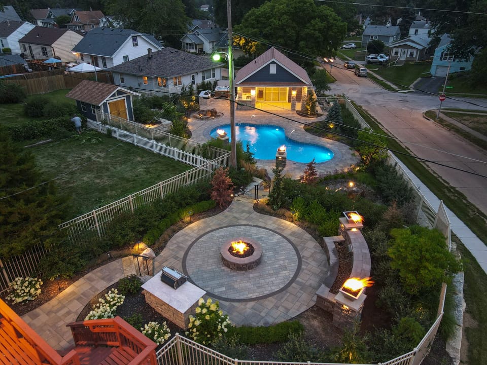 Aerial view of property featuring swimming pool, fire pit patio, and landscaped outdoor entertainment area in residential neighborhood setting.