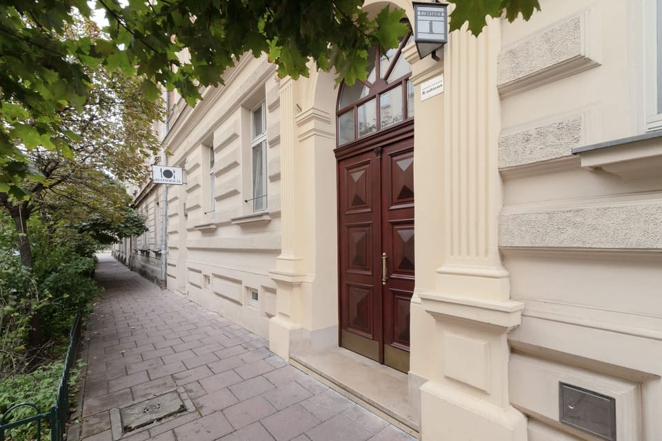 An exterior shot of a building entrance with ornate architectural details and greenery nearby.
