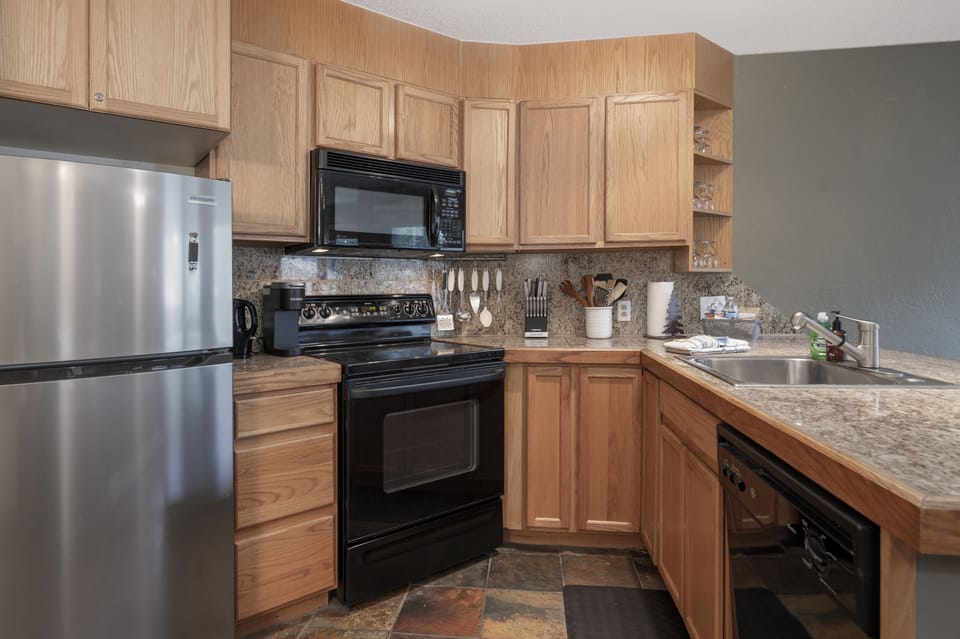 Kitchen with wooden cabinets, granite countertops, black appliances, stainless steel fridge, and all the essentials.