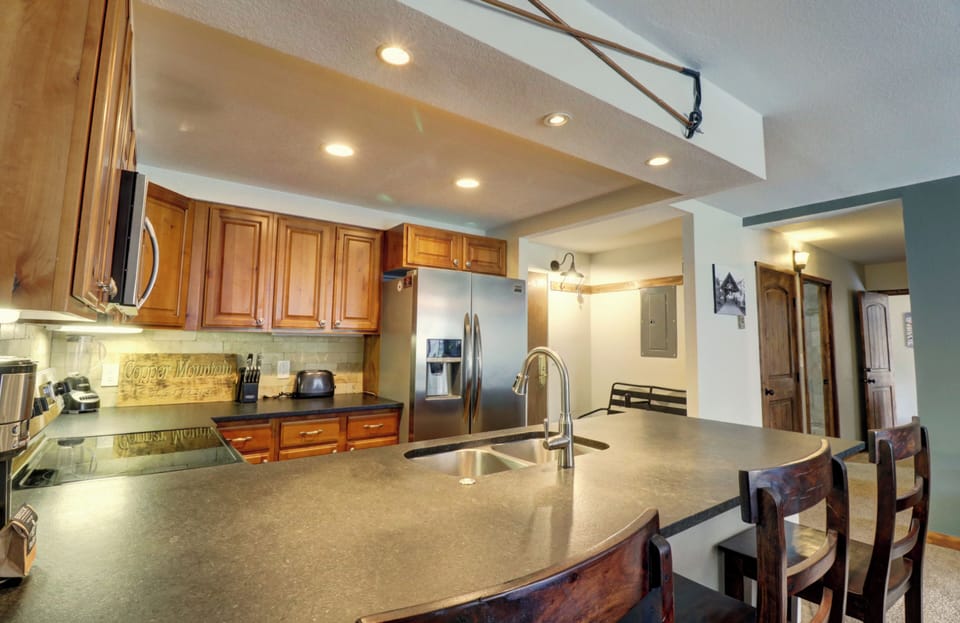 A modern kitchen with wooden cabinets, stainless steel appliances, and a large island with a sink and bar stools. Recessed lighting is installed in the ceiling.