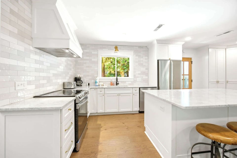 Warm oak planks anchor this airy kitchen, where shimmering subway tiles and cool stone surfaces meet a bright window view of lush greenery. Gold hardware adds a touch of shine to the clean cabinetry, creating a fresh space for morning coffee.