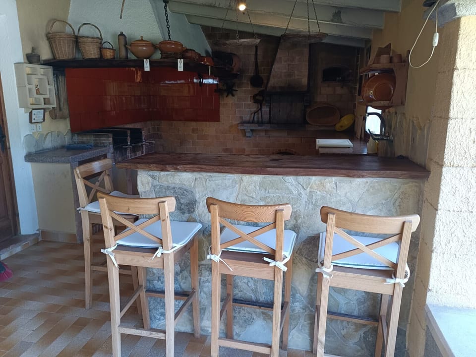 Outdoor kitchen with barbecue at Villa La Sort Llarga in Pollensa, Mallorca, featuring a stone bar and wooden stools, ideal for preparing and enjoying meals outdoors in an authentic Mediterranean atmosphere.