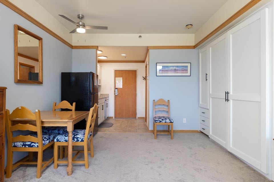 A compact dining area with a wooden table and chairs upholstered in blue fabric, a black refrigerator, and a door leading to a hallway. A large white cabinet is on the right wall.
