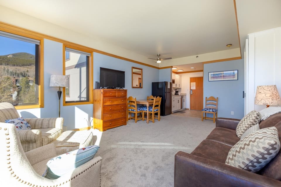 A brightly lit living area features a couch, armchair, wooden dresser, dining table with chairs, and wall-mounted TV. The adjacent kitchenette includes a refrigerator and cabinetry.