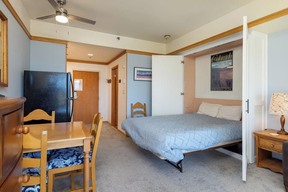 A compact studio apartment features a fold-out bed, a small dining table with chairs, a refrigerator, and a ceiling fan. The decor includes blue walls, wood trims, and a poster on the wall.