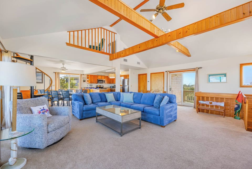 Relax in your spacious living room with soaring vaulted ceilings, exposed beams, and a vibrant blue sectional. Natural light pours through large windows, and the open layout flows into a fully-equipped kitchen and dining space—perfect for gathering with lo