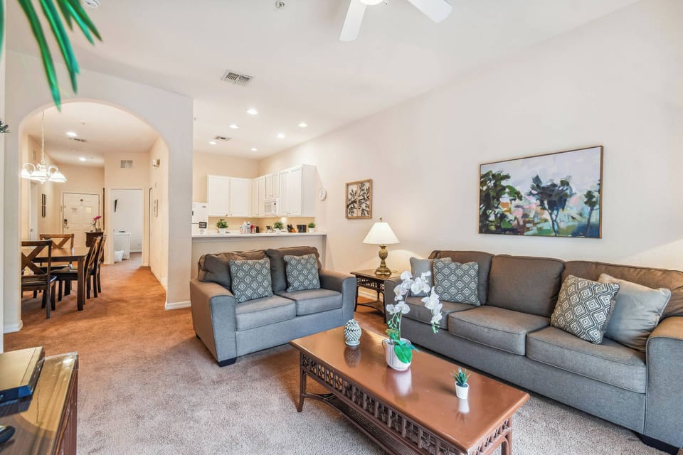 Welcome to a bright living area with soft beige walls and a cozy gray sofa, perfect for relaxation. The warm wood coffee table enhances the inviting atmosphere, while a view of the kitchen adds that comforting homey touch.
