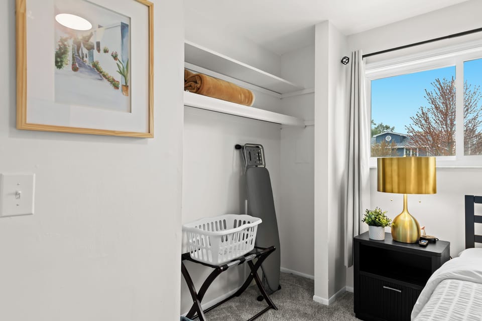 Cozy bedroom corner with open closet, ironing board, and a stylish gold lamp beside the bed.