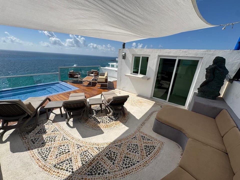 Terrace with stone mosaic floor, cushioned chaise lounges under shade sail, small pool on teak deck, and ocean views.