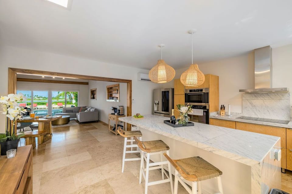 Picture yourself in this open, sunlit kitchen where marble countertops and warm wood cabinetry create a serene space for preparing meals while soaking in poolside views through the living room beyond.