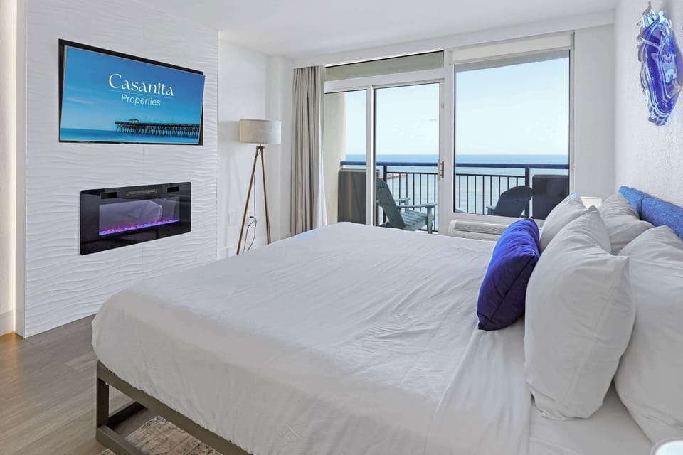 Relax in a bright Myrtle Beach studio with a cozy bed, wall-mounted TV, electric fireplace, and a comfortable coastal feel.