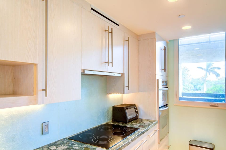 Light wood cabinets with long metal handles frame a black electric stovetop and green stone countertop in the kitchen.