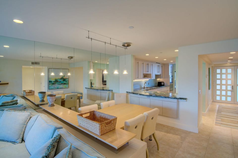 Dining table with cream-upholstered chairs sits beside a mirrored wall reflecting pendant lights and the kitchen’s granite countertop.