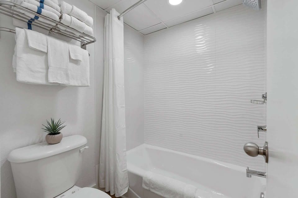A bright, modern bathroom with crisp white tiles, a relaxing bathtub, and plush towels awaits your stay! 
