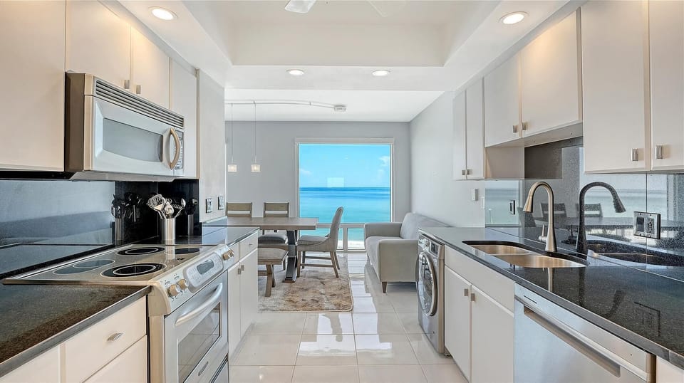 Galley kitchen with white cabinetry, black granite countertops, stainless steel appliances, and ocean view beyond dining area.