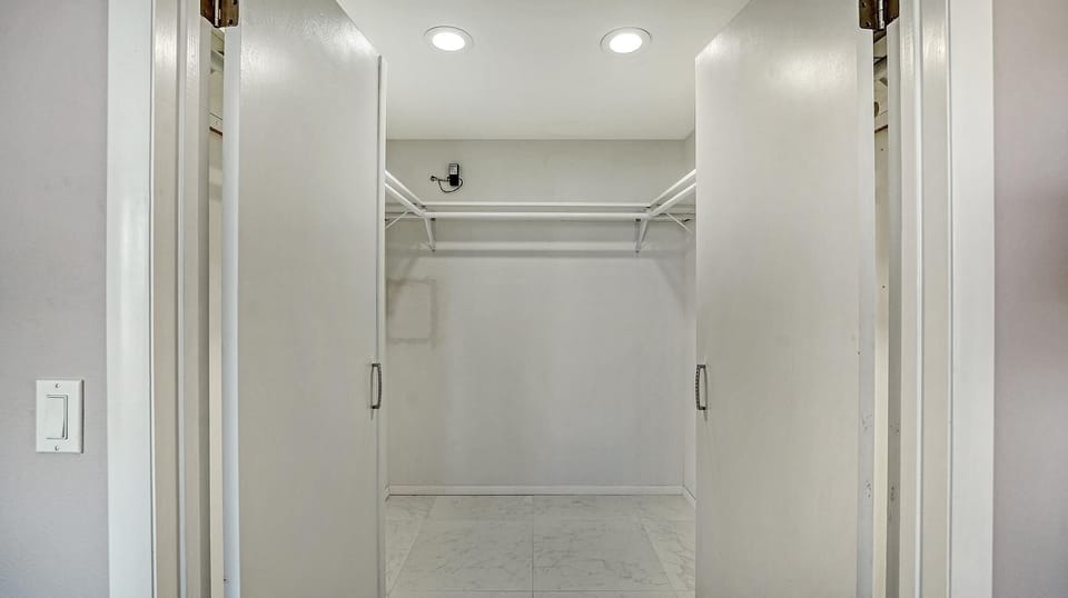 Walk-in closet with white double doors, metal hanging rods, and marble-look tile flooring under recessed lights.