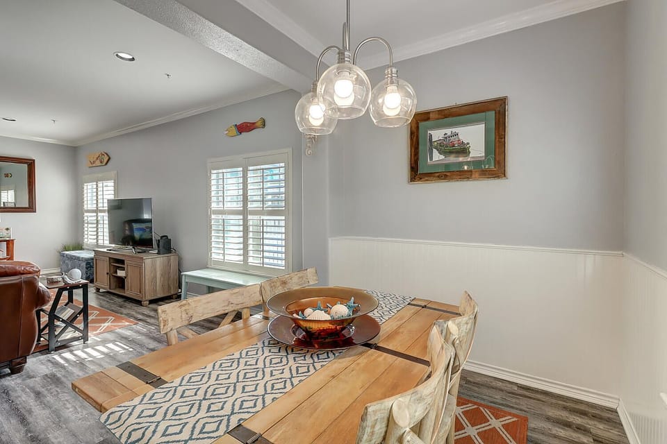 Your bright dining space features coastal charm with a rustic wooden table, pendant lighting, and an open view to the living area.