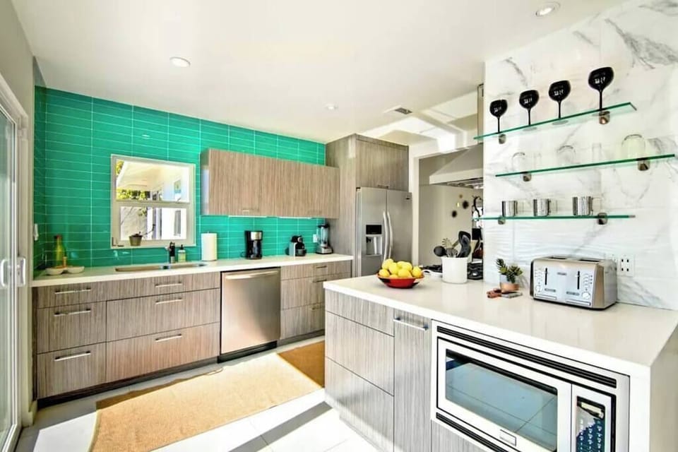 Sunlit kitchen with teal tile accent wall, stainless appliances, glass shelving, and generous island counter space