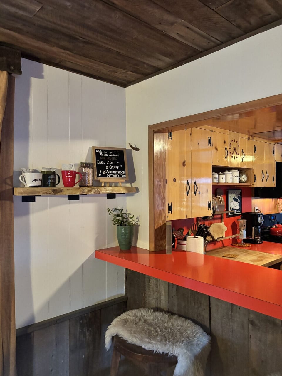 This inviting kitchen corner combines rustic wood with a bold red countertop. Shelves hold mugs and a welcome sign, while a potted plant and fluffy fur‑covered stool add cozy touches.